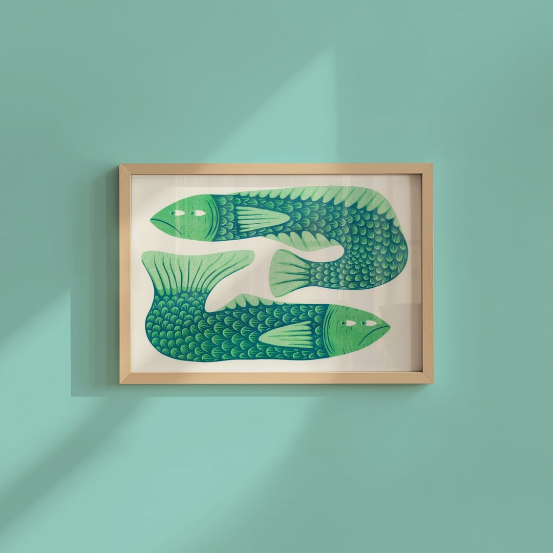 Framed wall art featuring two stylized green fish with detailed scales, displayed in a light wood frame on a mint green wall with soft diagonal shadows.