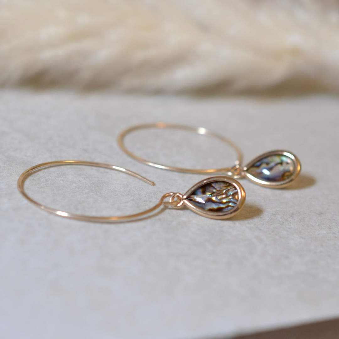Gold hoop earrings with teardrop-shaped paua shell dangle on a soft white background