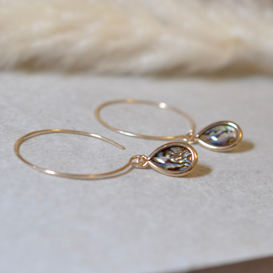 Gold hoop earrings with teardrop-shaped paua shell dangle on a soft white background