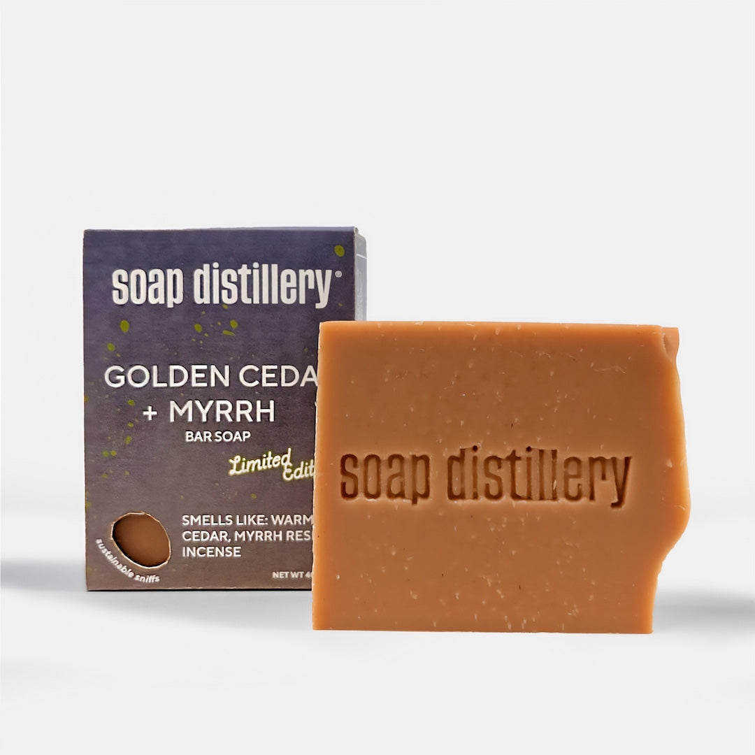 A bar of amber-brown soap, embossed with 'soap distillery', displayed next to its dark purple packaging box labeled 'GOLDEN CEDAR + MYRRH BAR SOAP' on a light gray background.