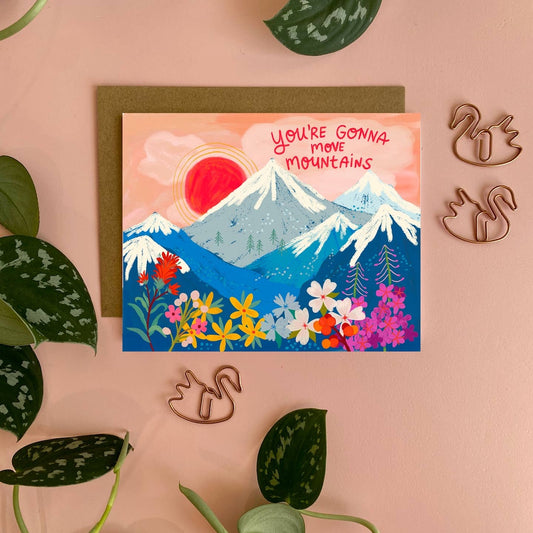 Card with mountain and floral design and text that says you're gonna move mountains on it on a pink background with decorative elements.