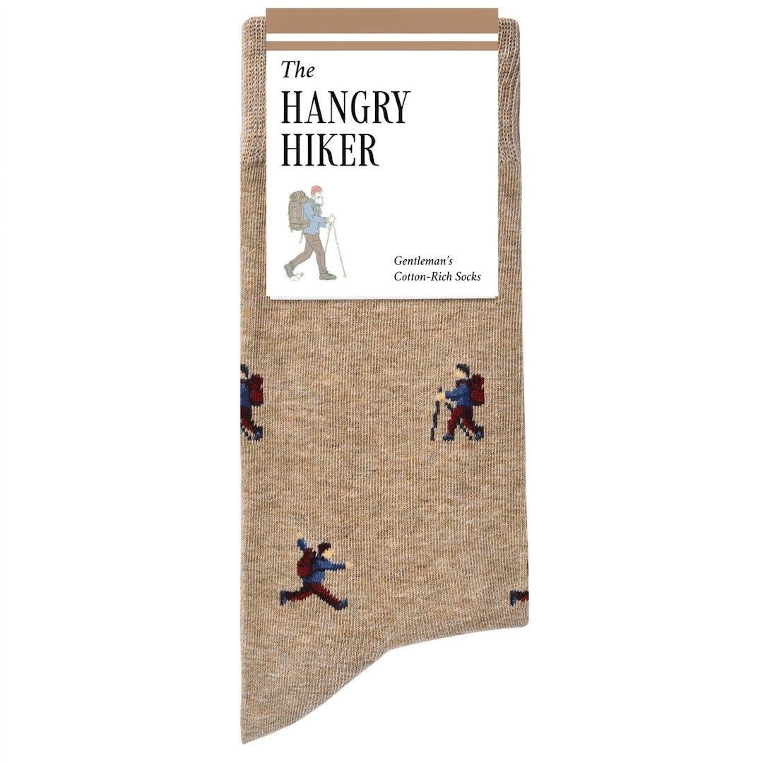 A pair of tan knit socks with a pattern of illustrated hikers, shown with a white paper header card that reads 'The HANGRY HIKER' on a plain white background.