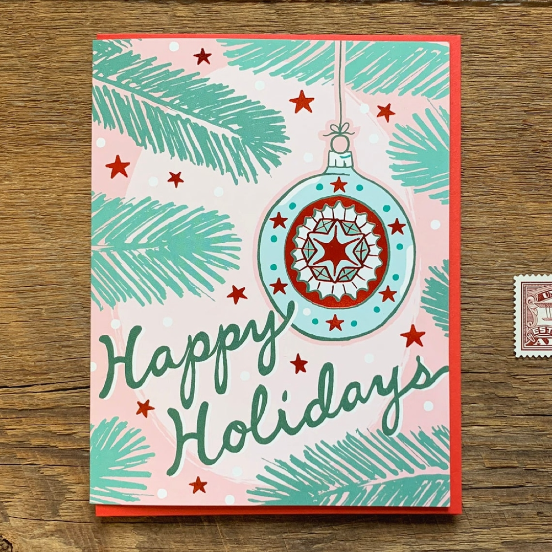 A square Christmas greeting card with a red border, featuring a pastel pink background, teal pine branches, red stars, and a large decorated ornament illustration. The card reads 'Happy Holidays' in dark teal script and is photographed on a wooden surface.
