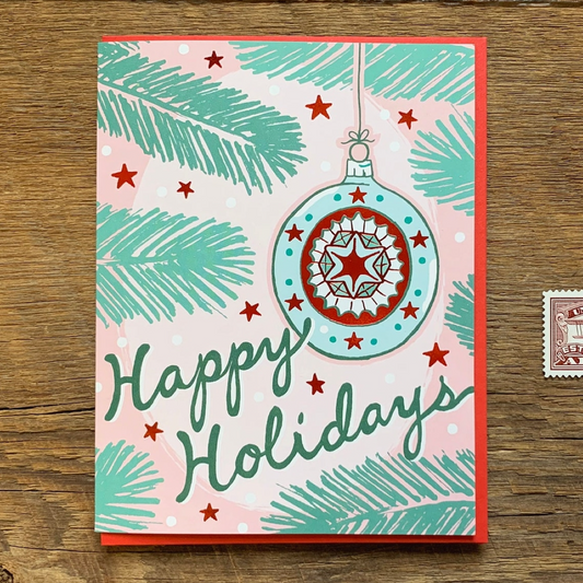 A square Christmas greeting card with a red border, featuring a pastel pink background, teal pine branches, red stars, and a large decorated ornament illustration. The card reads 'Happy Holidays' in dark teal script and is photographed on a wooden surface.