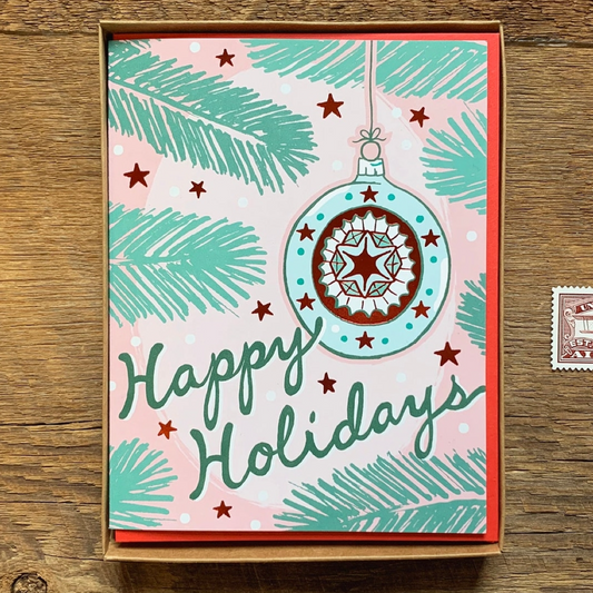 A square holiday greeting card in a kraft paper box, featuring a pink background with teal pine branches, red stars, and a large teal ornament illustration. The card reads 'Happy Holidays' in teal script and is photographed on a wooden surface.