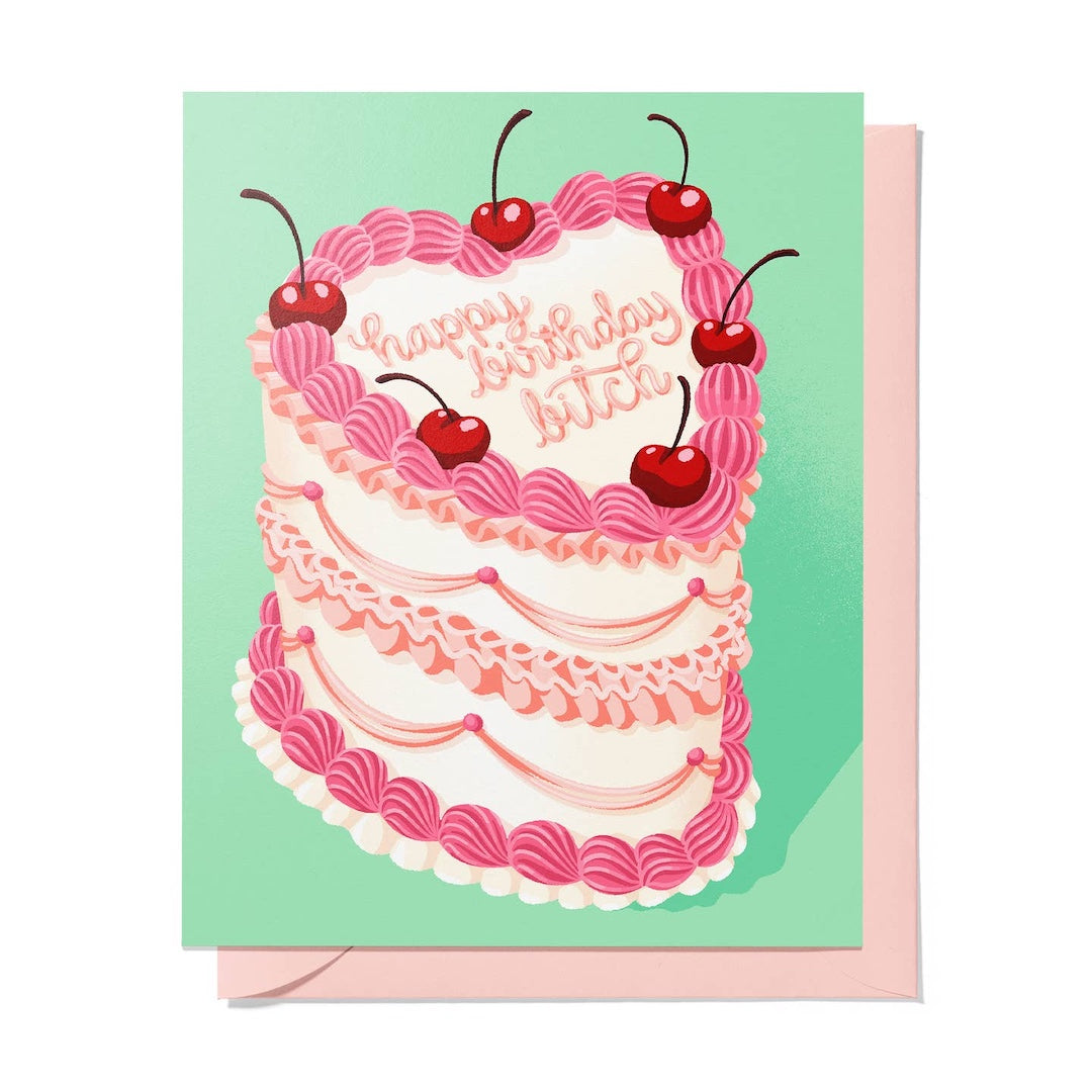 A square greeting card featuring an illustrated two-tier birthday cake with pink piped frosting, cherries, and the words 'happy birthday bitch,' shown with a light pink envelope behind it on a white background.