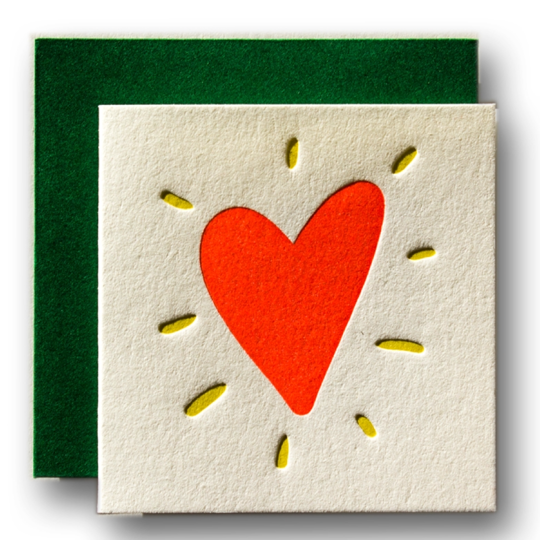 A square cream-colored greeting card with a textured surface, featuring a red heart in the center surrounded by small yellow felt rays, shown with a dark green felt backing card on a white background.