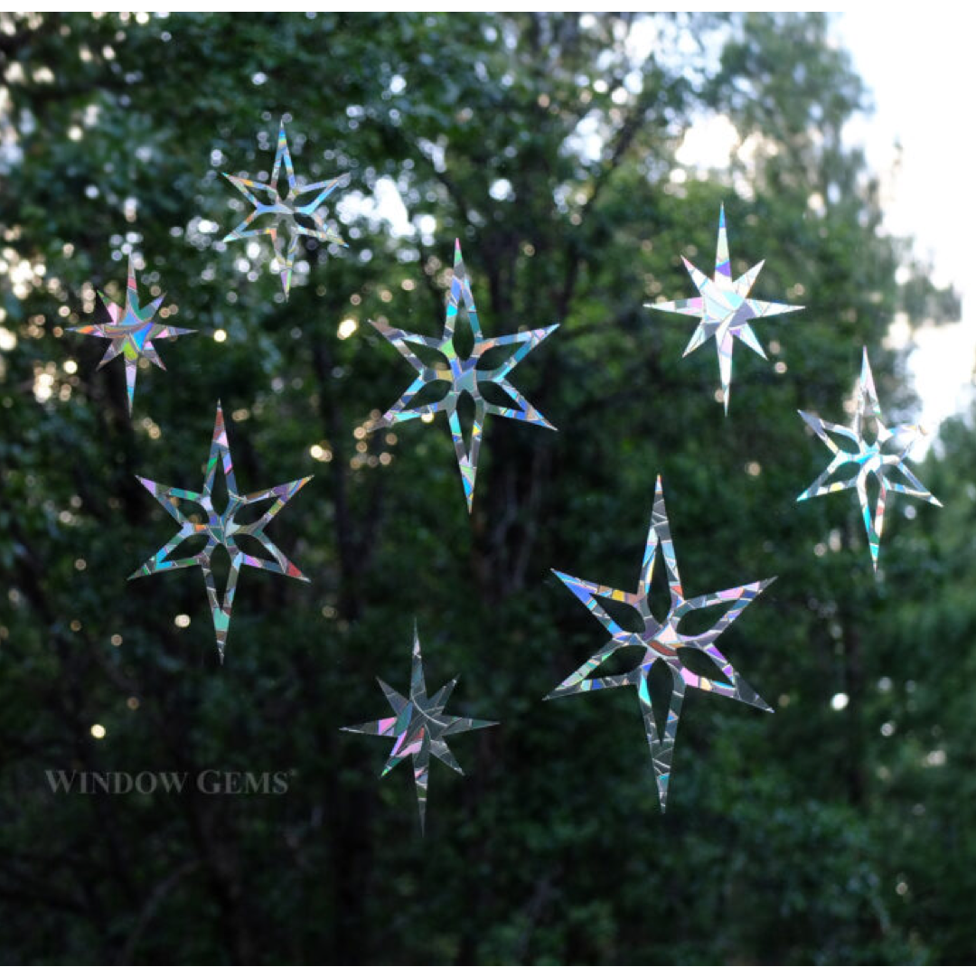 Seven iridescent star-shaped window gems are suspended against a blurred background of green trees and bright sunlight. The brand name 'WINDOW GEMS' is printed in white text in the lower-left corner.
