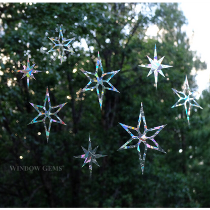 Seven iridescent star-shaped window gems are suspended against a blurred background of green trees and bright sunlight. The brand name 'WINDOW GEMS' is printed in white text in the lower-left corner.