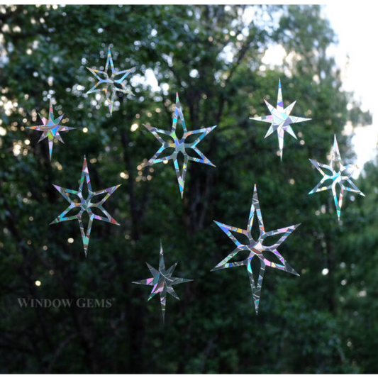 Seven iridescent star-shaped window gems are suspended against a blurred background of green trees and bright sunlight. The brand name 'WINDOW GEMS' is printed in white text in the lower-left corner.