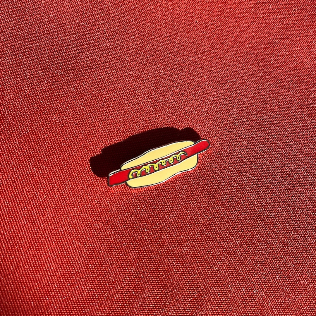 A small enamel pin shaped like a hot dog bun with a red hot dog and mustard, placed on a textured red fabric background.