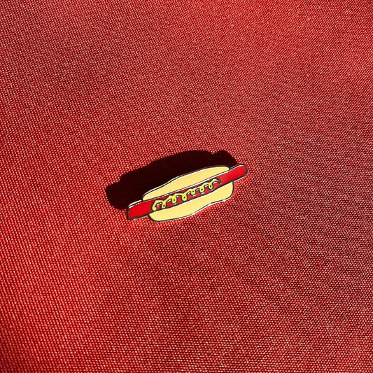 A small enamel pin shaped like a hot dog bun with a red hot dog and mustard, placed on a textured red fabric background.