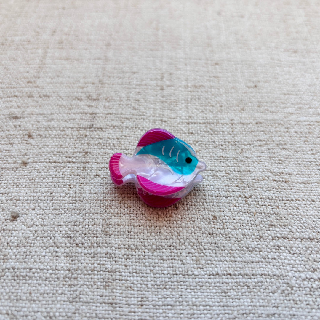 A small fish-shaped hair clip with a pink and teal pattern, placed on a beige woven fabric background.