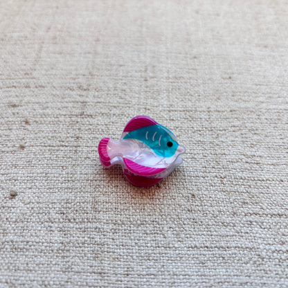 A small fish-shaped hair clip with a pink and teal pattern, placed on a beige woven fabric background.