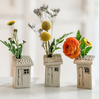 Three small ceramic vases painted to look like houses, each containing different arrangements of flowers on a white background.