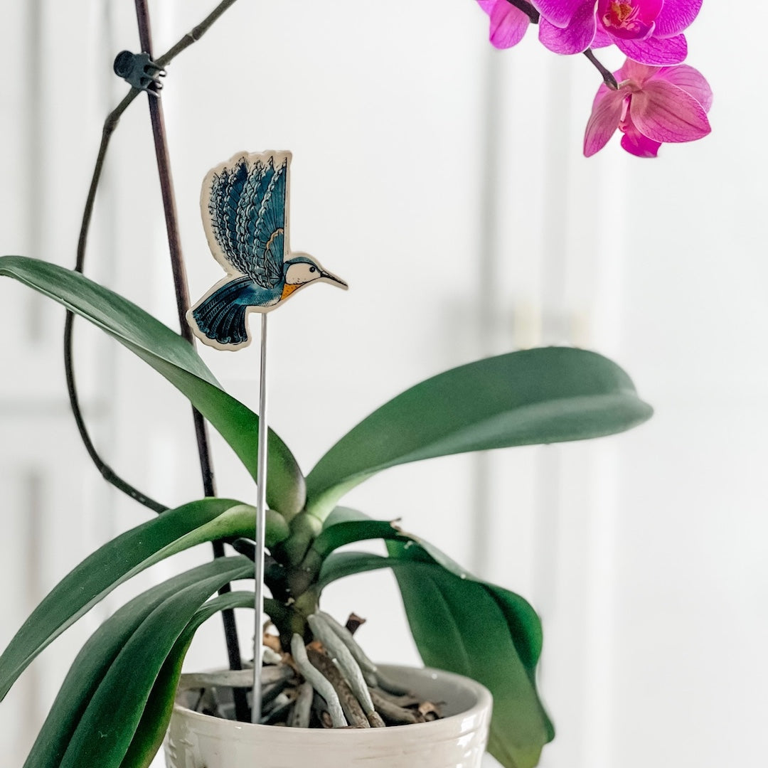Decorative hummingbird figurine on a stick in a potted plant with pink flowers in the background