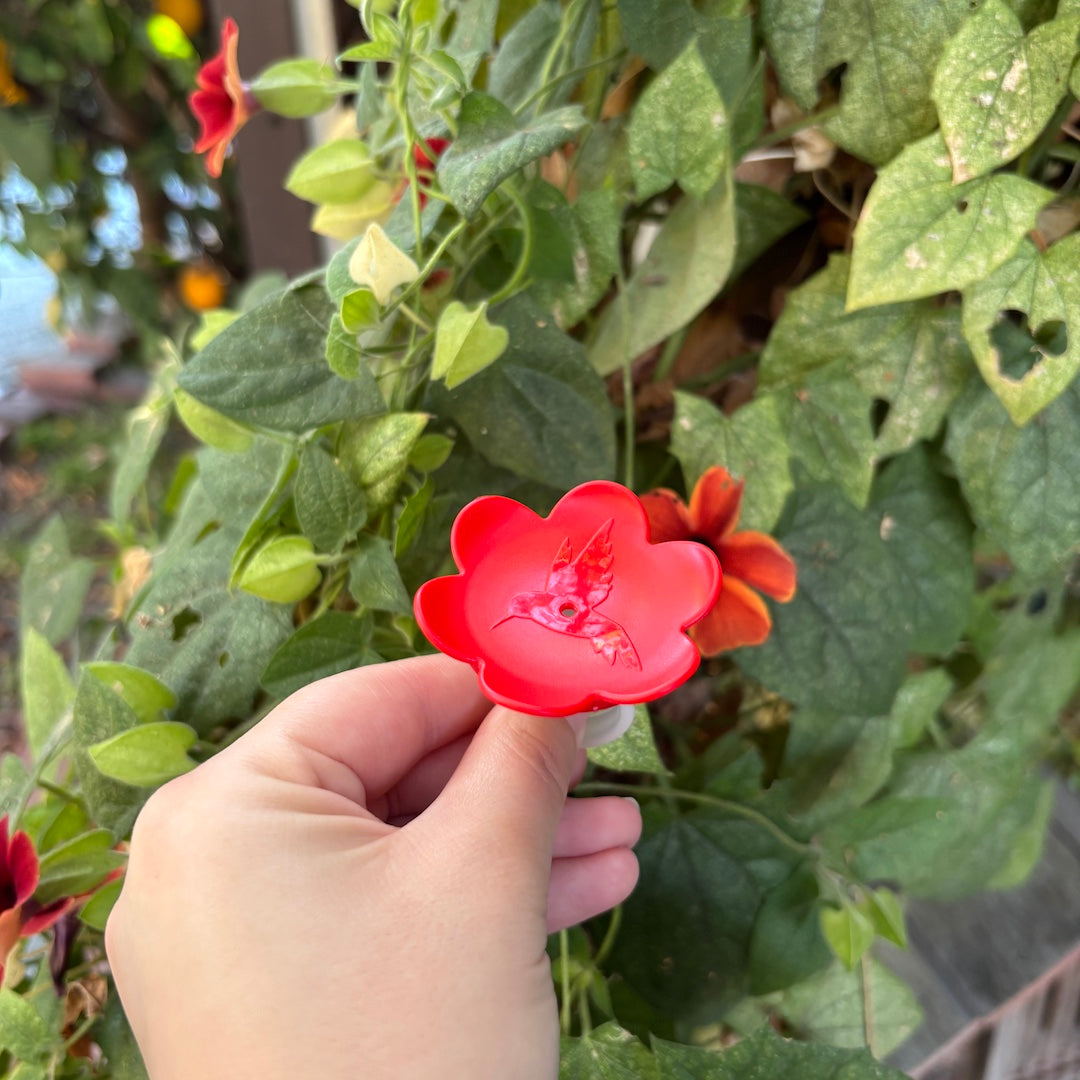A hand holding a bright red plastic flower-shaped hummingbird feeder ring with a hummingbird ornament, set against a background of green foliage and orange flowers.