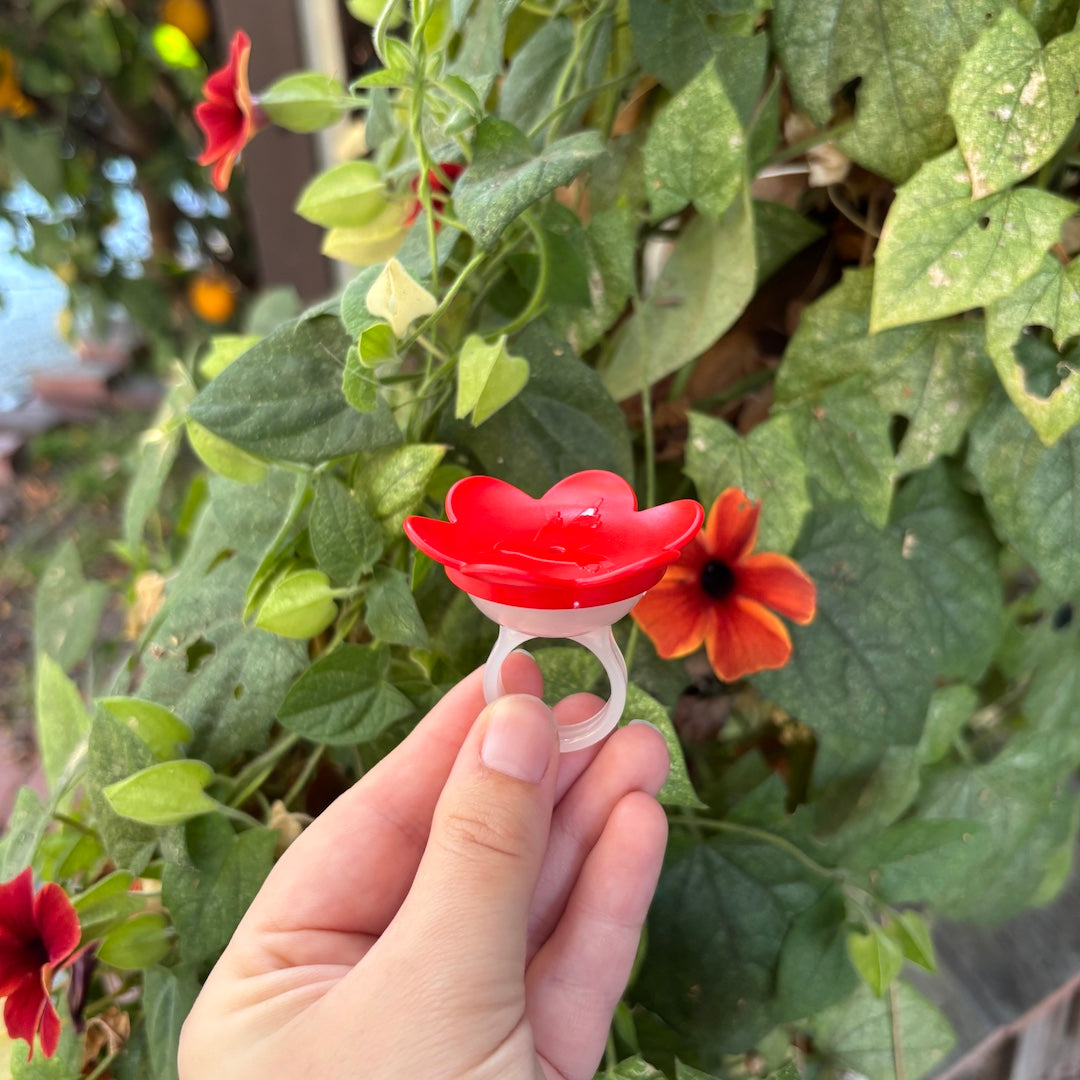 A hand holds a red flower-shaped ring holder outdoors in front of green foliage and orange flowers.