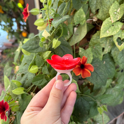 A hand holds a red flower-shaped ring holder outdoors in front of green foliage and orange flowers.