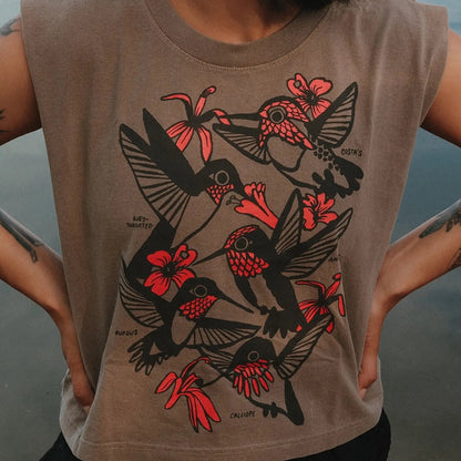 A sleeveless taupe-brown crop top featuring a large graphic print of black and red hummingbirds and flowers, worn by a model with arms visible.