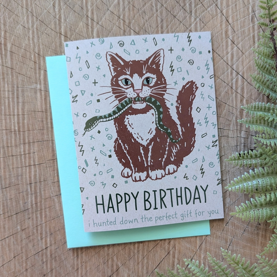 A square greeting card featuring a brown tabby cat holding a green snake in its paws, with the text 'HAPPY BIRTHDAY i hunted down the perfect gift for you' printed below. The card is placed on top of a light blue envelope on a rustic wood surface with green fern leaves at the side.