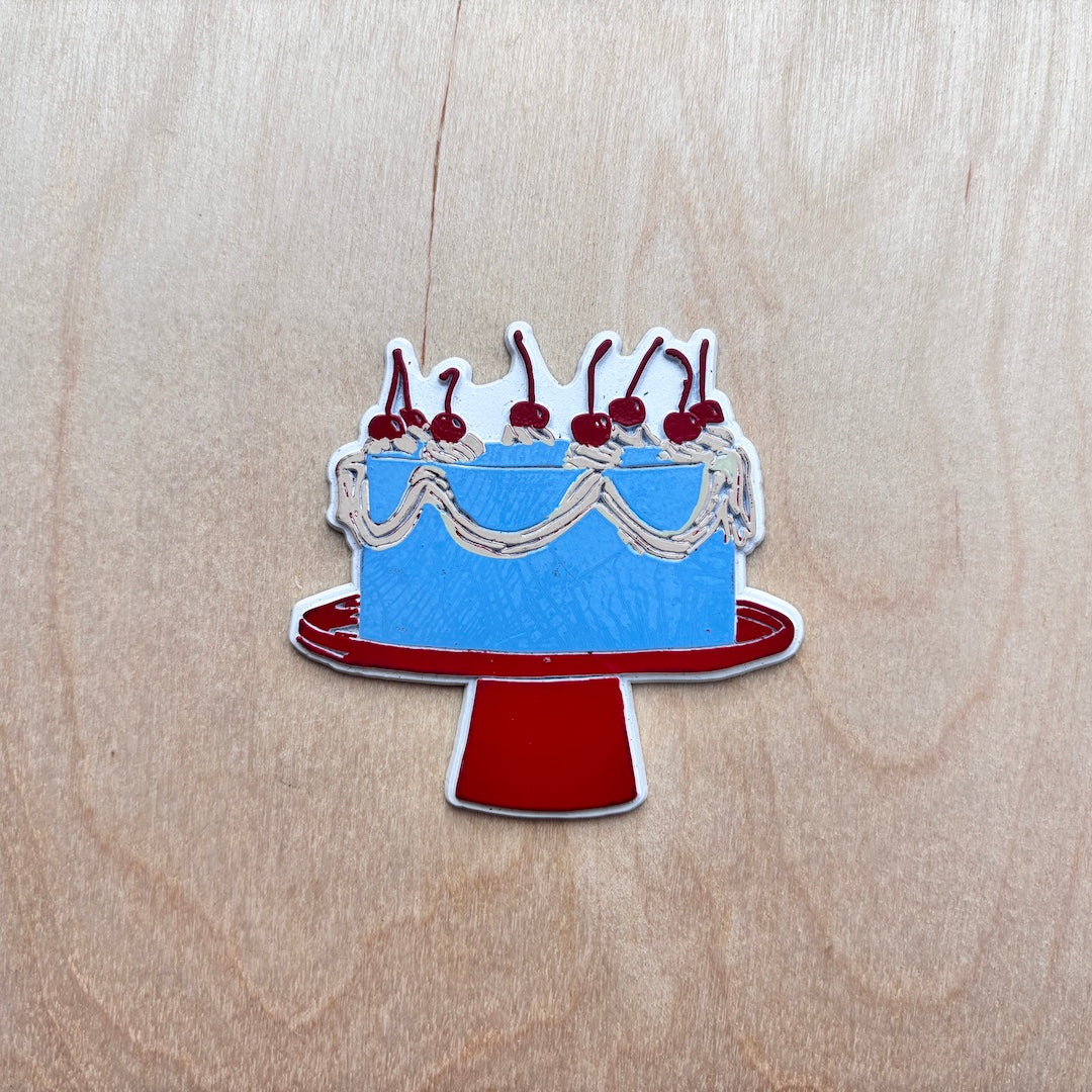 A fridge magnet shaped like a blue cake on a red stand, decorated with white frosting, cream swirls, and red cherries, placed on a light wood background.