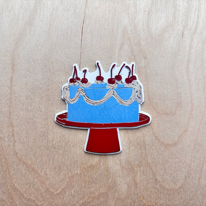 A fridge magnet shaped like a blue cake on a red stand, decorated with white frosting, cream swirls, and red cherries, placed on a light wood background.