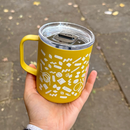 A hand holding a yellow insulated tumbler with a clear lid and white doodle-style pasta graphics, photographed outdoors on a paved walkway.