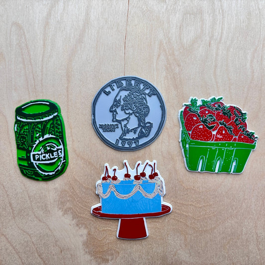 Four novelty fridge magnets shaped like a green pickle jar, a quarter, a blue cake with cherries on a red stand, and a green basket of red strawberries, arranged on a light wood background.