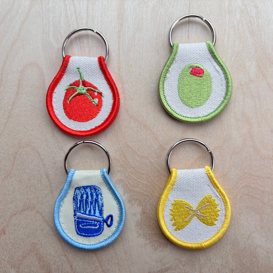 Four teardrop-shaped embroidered keychains on a light wood background. Each keychain has a white center, a colorful embroidered food or object design, and a silver metal key ring.