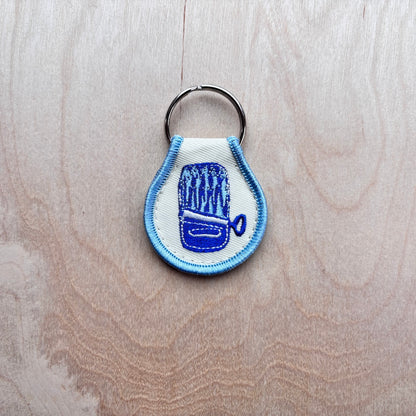 A small embroidered keychain charm with a silver metal key ring, featuring a blue and white design of an open can of sardines, placed on a light wood grain background.