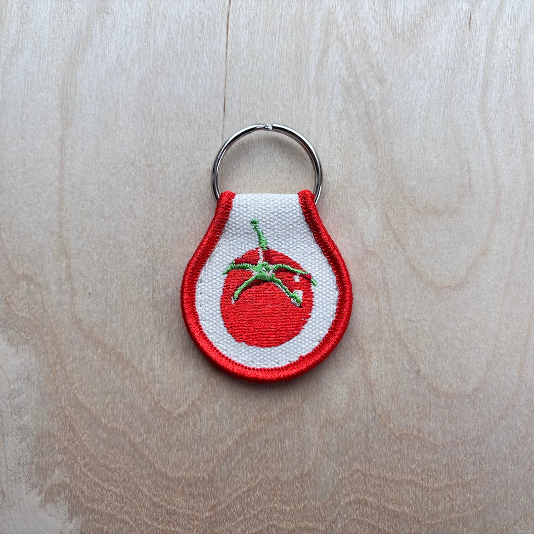 An embroidered tomato keychain charm with a red border, white center, red tomato motif with green stem, attached to a silver metal key ring, placed on a light wood surface.