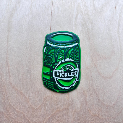 A green fridge magnet shaped like a pickle jar with a white circular logo reading 'PICKLES', placed on a light wood grain background.