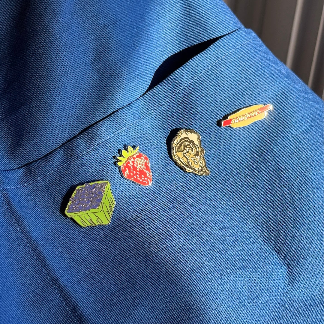 Four colorful enamel pins shaped like a strawberry, a hot dog, a green box, and a gold shell, displayed on a blue denim jacket.