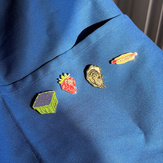 Four colorful enamel pins shaped like a strawberry, a hot dog, a green box, and a gold shell, displayed on a blue denim jacket.