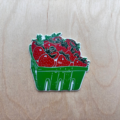 A fridge magnet depicting a green plastic berry basket filled with red strawberries, placed on a light wood grain surface.