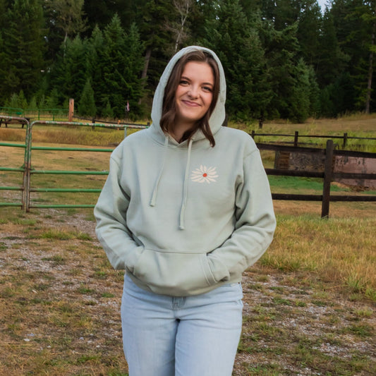A woman wearing a sage green hoodie with a white daisy graphic on the chest, standing outdoors in front of a wooden fence and trees.