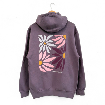 A dark purple hoodie with a large floral graphic on the back, featuring pink, white, and dark purple flowers with yellow centers, hanging on a wooden hanger against a white background.