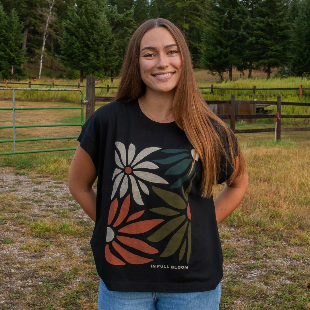 A smiling young woman stands outdoors in a grassy field with trees and a fence behind her, wearing a black graphic T-shirt with large floral designs in white, rust orange, and olive green, plus blue jeans. The shirt has small white text near the lower hem reading 'IN FULL BLOOM'.