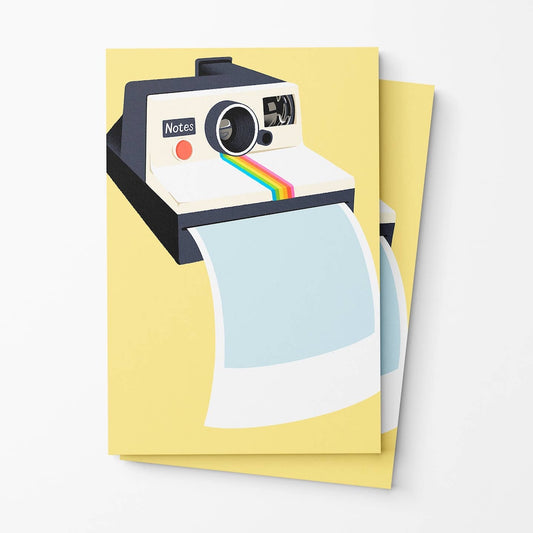 A yellow Polaroid-style instant camera illustration printed on a notepad, with two overlapping cards shown against a light gray background. The front card features a pale blue and white polaroid photo strip graphic.
