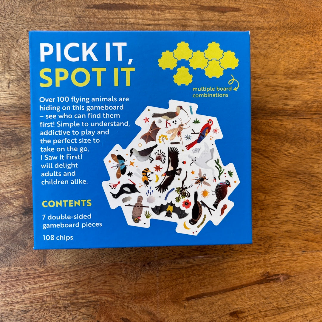 The back of the packaging box for the board game 'I saw it first flying' lies flat on a wooden tabletop. The box is bright blue with yellow and white text, featuring an illustration of various animal-themed game pieces.