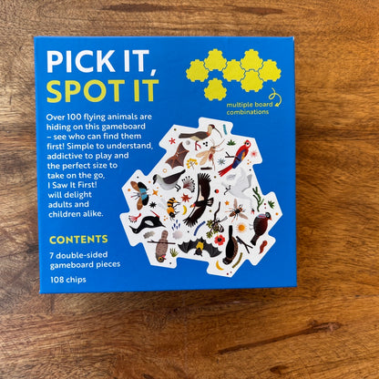 The back of the packaging box for the board game 'I saw it first flying' lies flat on a wooden tabletop. The box is bright blue with yellow and white text, featuring an illustration of various animal-themed game pieces.