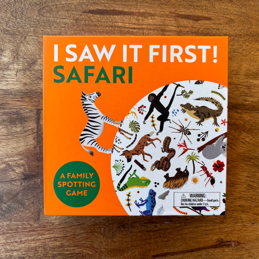 The board game box for 'I Saw It First! Safari' is shown flat on a wooden tabletop. The box features a bright orange background with white and green text, an illustration of a zebra, and a white circular section displaying various safari animals in colorful illustrations.