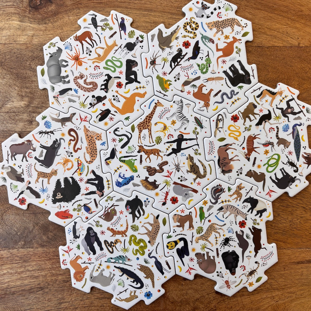 A set of interlocking animal-themed puzzle pieces arranged in a circular pattern on a wooden tabletop. Each piece features colorful illustrations of different animals, including giraffes, zebras, lions, bears, snakes, and birds, on a white background.