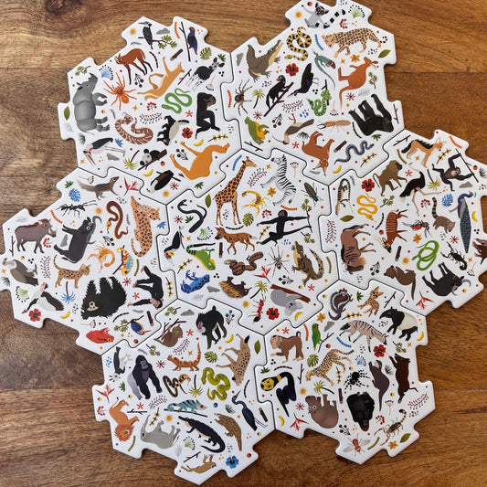 A set of interlocking animal-themed puzzle pieces arranged in a circular pattern on a wooden tabletop. Each piece features colorful illustrations of different animals, including giraffes, zebras, lions, bears, snakes, and birds, on a white background.