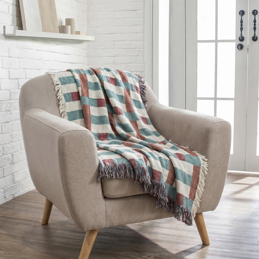 A beige upholstered armchair with light wood legs in a bright room, draped with a fringed throw blanket featuring a muted plaid pattern in cream, teal, and terracotta.