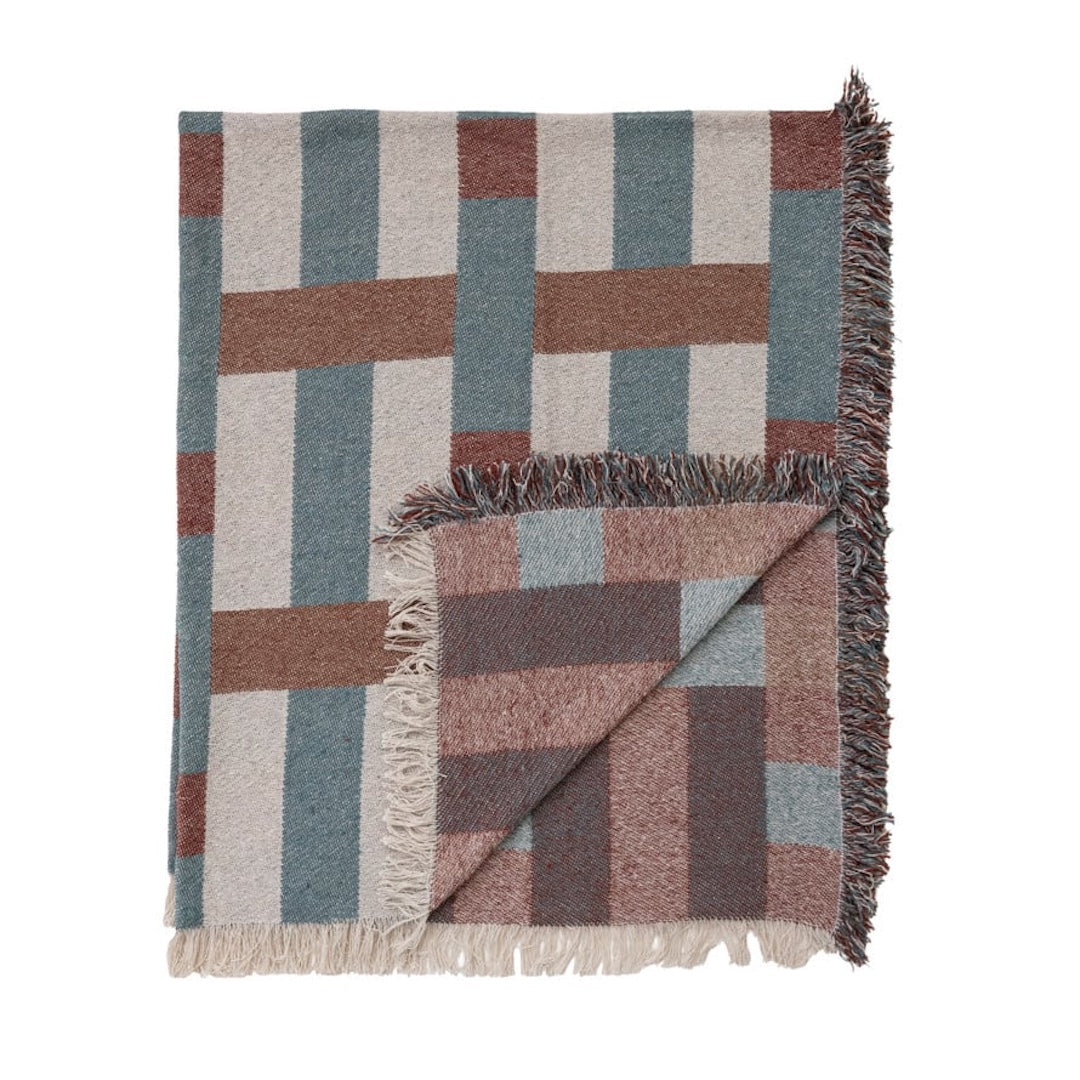 A woven throw blanket with a geometric block pattern in muted tones of slate blue, terracotta, dusty rose, and cream, featuring fringed edges and shown folded on a white background.