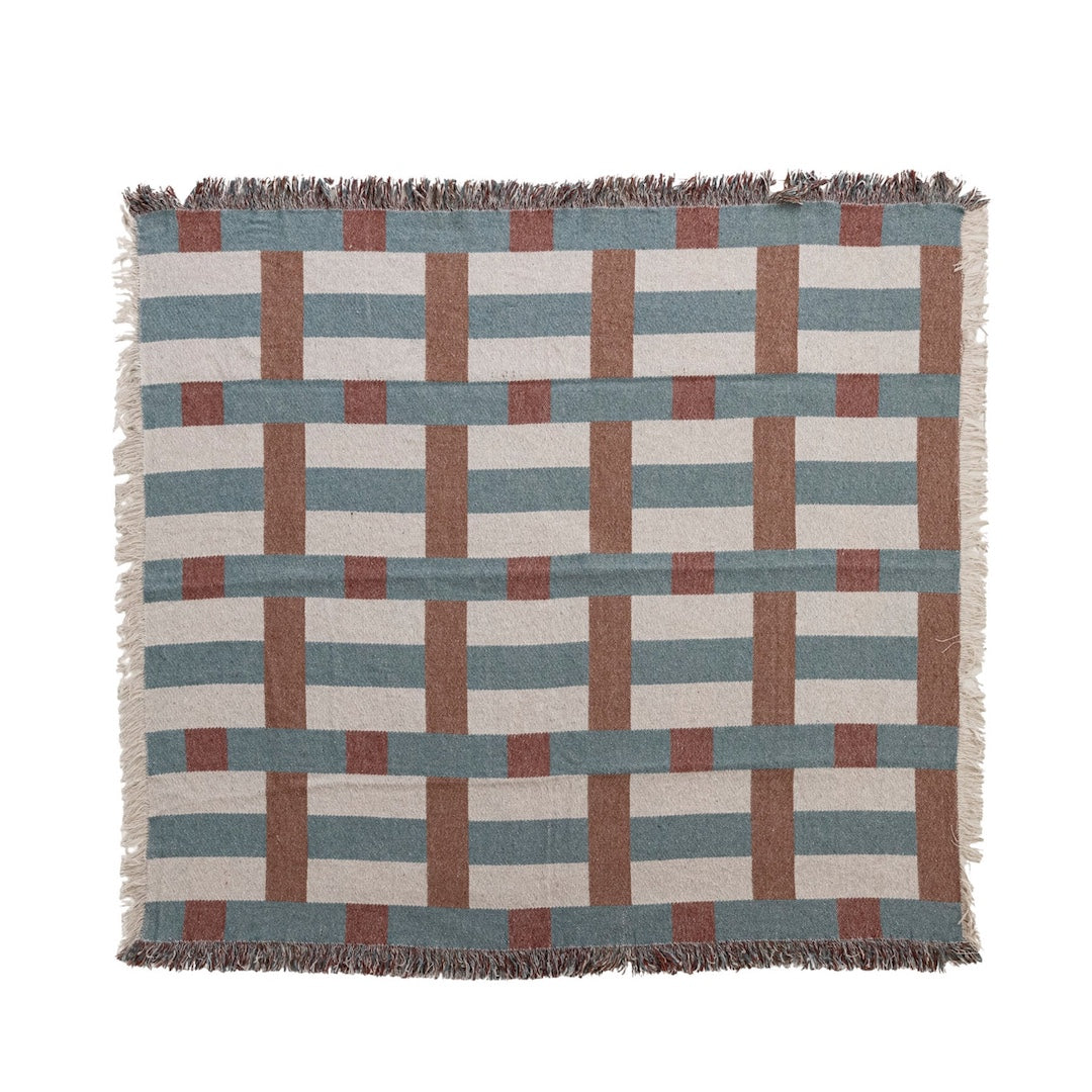A woven throw blanket with a geometric block pattern in muted tones of slate blue, terracotta, dusty rose, and cream, featuring fringed edges