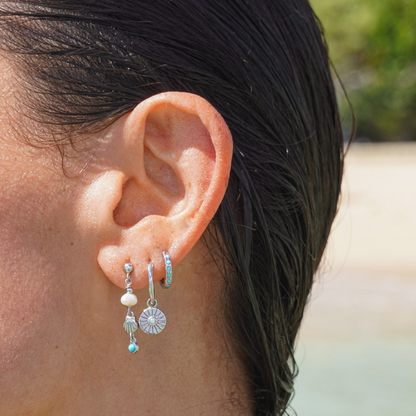 A close-up of a person's ear wearing multiple silver-toned dangle earrings, including a small hoop, a beaded drop, a shell charm, and a sunburst disc charm, with turquoise blue beads and a white pearl.