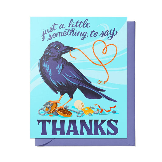 A greeting card with a light blue front featuring an illustrated dark blue raven holding an orange rope that forms a heart shape, with the text 'just a little something to say' above and 'THANKS' below. A lavender envelope is visible behind the card.